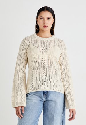 Crème-kleurige gebreide trui met een gestreept patroon, bootnek en uitlopende mouwen. Draagt over lichtblauwe high-waisted jeans.
