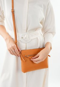 Piccola borsa a tracolla in pelle arancione con finitura testurizzata, tasca anteriore con zip e tracolla regolabile, tenuta da una persona in un outfit bianco.