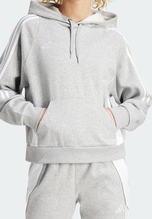 Henkilö, joka pukeutunut vaaleanharmaaseen Adidas-huppariin ja siihen sopiviin collegehousuihin, joissa valkoiset raidat, käsivarret hupun vetoketjutaskussa.