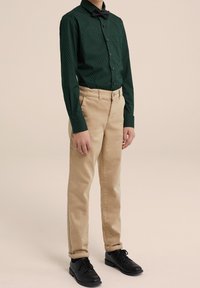 Mörkgrön, långärmad skjorta med vita prickar, i kombination med ljusbeige byxor och svarta skor. Fluga ger en formell prägel.