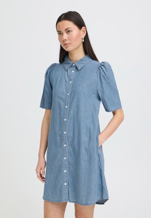 Nő rövid ujjú, gombos, kék farmeranyagból készült ruhát visel, amelynek gallérja és oldalzsebei vannak, egy egyszínű fehér háttér előtt áll.