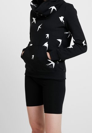 Schwarzer Hoodie mit weißen Vogelmotiven und Fronttasche, getragen mit schwarzen Radshorts bei einer Person, die vor einem einfarbigen Hintergrund steht.