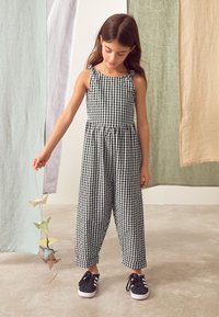 Tuta a quadretti bianca e nera con corpetto aderente, gambe larghe e spalline regolabili. Bambino che tiene in mano figure di origami, indossando scarpe da ginnastica nere.