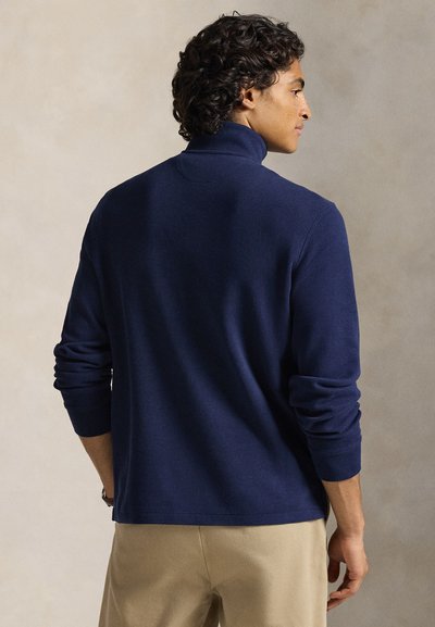 Göndör hajú férfi, sötétkék hosszú ujjú garbó pulóvert és bézs nadrágot visel, semleges háttér előtt áll.