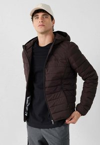 Giacca imbottita marrone scuro con cappuccio, caratterizzata da una zip frontale, texture trapuntata e interno nero. Indossata sopra una t-shirt nera.