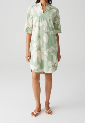 Mujer con un vestido suelto de longitud hasta la rodilla, de color verde claro con grandes patrones florales blancos y mangas cortas, combinado con sandalias blancas.