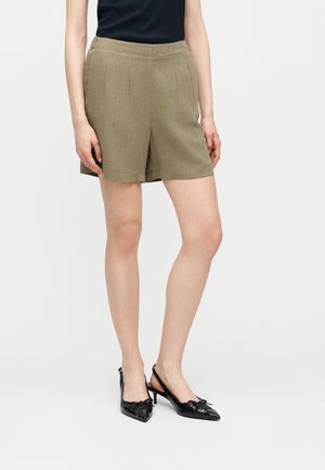 Kvinde iført olivengrønne knælange shorts, sort ærmeløs top og sorte slingback-hæle med spidst tåparti og sløjfedetalje, stående mod en hvid baggrund.