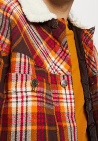 Geruit jasje met bruine, oranje en rode kleuren; heeft een pluizige kraag, drukknopen en een warme stoftextuur. Draagt over een mosterdkleurig overhemd.