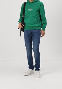 Zielona bluza z kapturem z centralnym logo, ciemnoniebieskie dżinsy i białe trampki. Bluza ma przednią kieszeń i sznurek do regulacji, luźny krój.