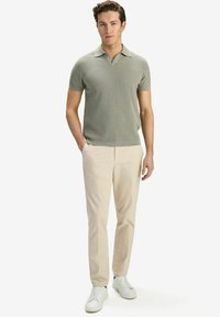 Ljusgrön, kortärmad polotröja med stickad struktur; kombinerad med krämfärgade chinos och vita sneakers, stående framför en neutral bakgrund.