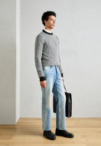 Grijs gemusterde trui met zwarte accenten, lichtblauwe jeans en zwarte laarzen, met een zwarte tote bag in de hand, staande op een houten vloer.