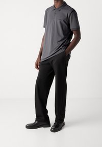 Polo shirt gris foncé avec un col, des manches courtes et un logo discret. Associé à un pantalon noir et des chaussures noires, mettant en valeur une coupe ajustée.