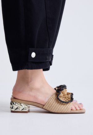 Pie que lleva una mule beige tejida con volante rematado en negro y tacón corto metálico de bloque, combinado con pantalones negros remangados al tobillo.