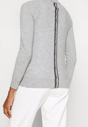 Person iført en lysegrå sweater med en lodret sort, hvid og rød logostribe ned ad ryggen, kombineret med hvide bukser.