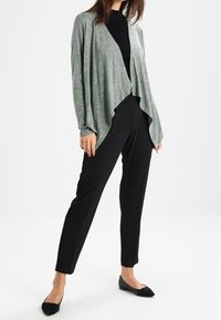 Světle zelený kardigan s otevřeným předním dílem, dlouhými rukávy a splývavým střihem, zkombinovaný s černými upnutými kalhotami a botami.