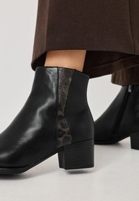 Botín negro de piel hasta el tobillo con un panel lateral texturizado y con patrón, y un tacón cuadrado. Superficie lisa, detalle sutil de la marca en el tacón.