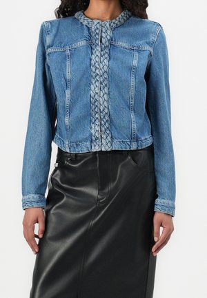 Femme portant une veste en jean bleue avec une bordure tressée à l'avant et aux poignets, associée à une jupe en cuir noire.