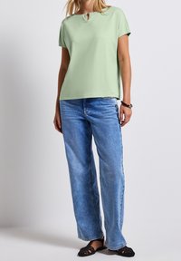 Ljusgrön kortärmad topp med rund hals och nyckelhålsdetalj, ihop med vida ljusblå jeans och svarta slip-on skor.