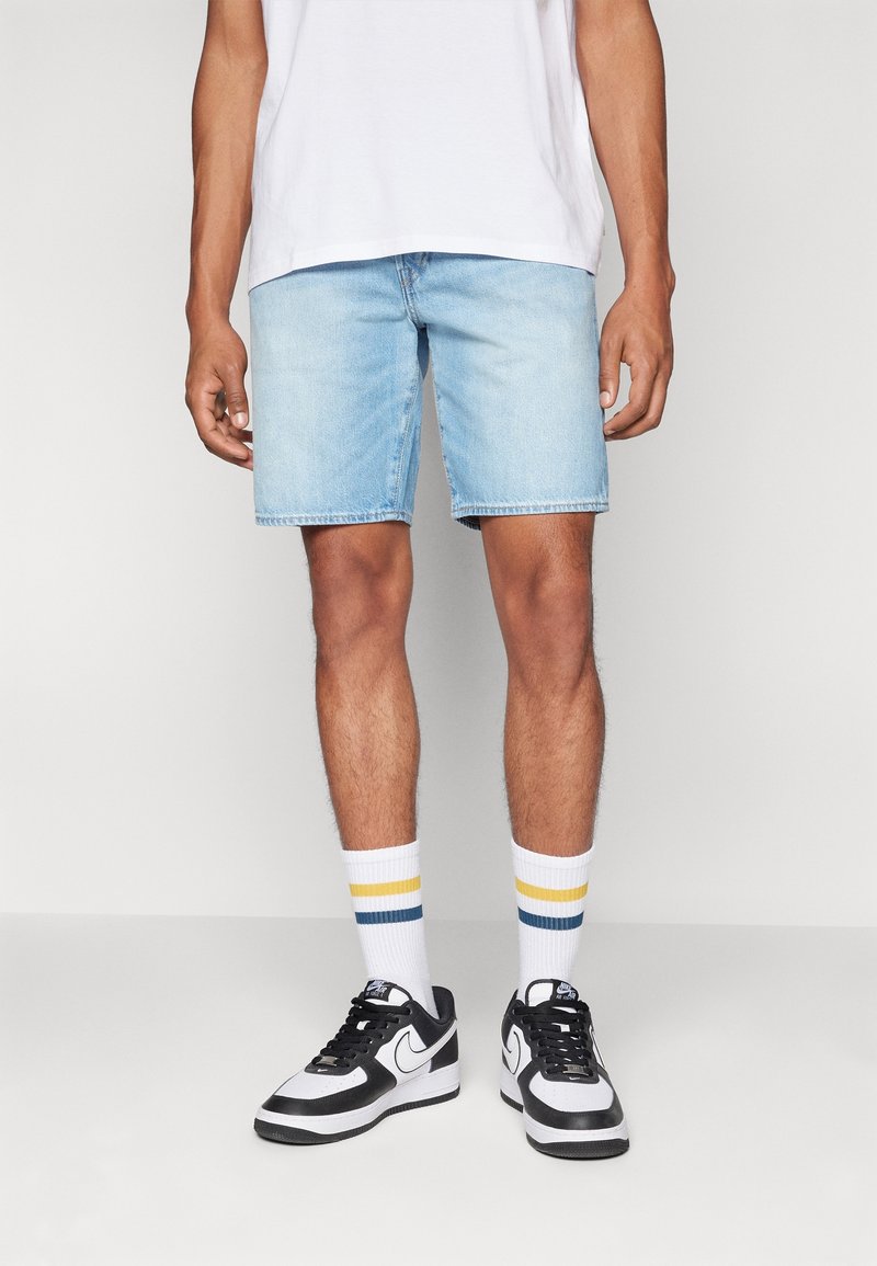 Shorts en denim bleu clair à coupe décontractée, portés avec des chaussettes blanches à rayures jaunes et bleues, associés à des baskets noires et blanches.