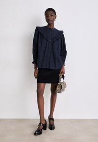 Marineblaue Rüschbluse mit langen Ärmeln, Knopfverschluss und strukturiertem Stoff; kombiniert mit einem schwarzen Cord-Rock und schwarzen High Heels. Handtasche im Schlangenprint.
