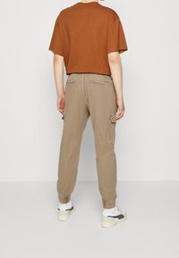 Hnědé oversized tričko, béžové cargo kalhoty s nařasenými kotníky, s bočními kapsami a uvolněným střihem, doplněné bílými teniskami.
