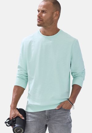 Mann mit kurzen Haaren, trägt ein hellblaues langärmeliges Hemd und graue Jeans, hält einen schwarzen tragbaren Lautsprecher in der linken Hand.