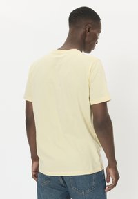 Mann, der von hinten blickt, trägt ein blassgelbes T-Shirt mit kurzen Ärmeln und blaue Jeans vor einem schlichten weißen Hintergrund.