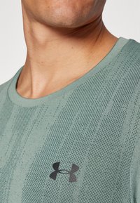 Világoszöld, rövid ujjú póló kerek nyakkivágással, texturált mintával és fekete Under Armour logóval a mellkasán.