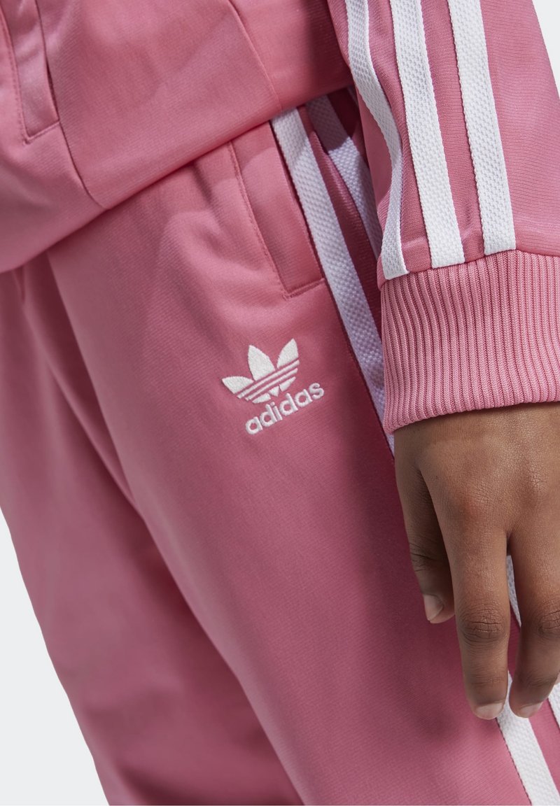 Różowy dres z białymi paskami, z prążkowanymi mankietami i wyszywanym logo Adidas na biodrze, charakteryzujący się miękką, gładką fakturą.