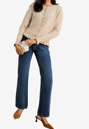 Femme portant un cardigan beige à volants boutonné, un jean droit bleu foncé, des chaussures marron à talons bas, tenant une petite pochette beige.