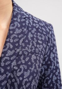 Námořnický blazer s texturovaným leopardím vzorem v bílé barvě. Má klopový límec, jednu kapsu a strukturovaný střih. Hladká tkanina.