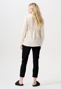 Hellbeige strukturierte Bluse mit langen Ärmeln und tailliertem Schnitt, kombiniert mit schwarzen Slim-Fit-Hosen und schwarzen Rutsch-Sandalen.