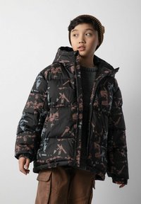 Dziecko w wzorzystej czarnej kurtce puchowej, brązowych spodniach cargo i brązowej czapce w paski, patrzy na bok na tle jednolitego tła.