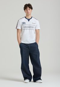 Hvid kortærmet trøje med sort "UMBRO" logo og marineblå accenter; parret med løse mørkeblå denimbukser og hvide sportsko.