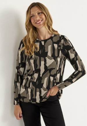 Kvinde med langt blond hår smiler iført en sort, beige og grå abstrakt mønstret langærmet top og sorte bukser, stående mod en hvid væg.