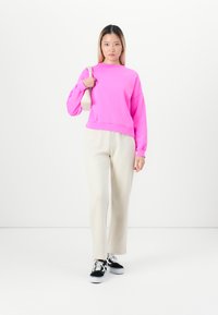 Young woman standing in beige pants, bright pink sweatshirt, black-and-white sneakers, holding white shoulder bag against white background.