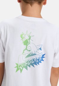 Dítě v bílém tričku s zeleným a modrým logem Quiksilver, na kterém je stylizovaná postava s hvězdou a surfovacími prknem na zádech.