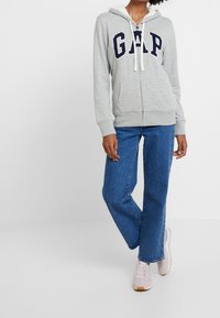 Persona vestida con una sudadera gris con cremallera de GAP y cordones blancos, jeans de corte recto azul y zapatillas de deporte rosa claro, de pie contra un fondo blanco.