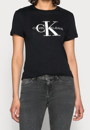 Femme portant un t-shirt noir Calvin Klein Jeans avec un grand logo blanc "CK" et un jean noir délavé, debout avec les mains détendues le long du corps.