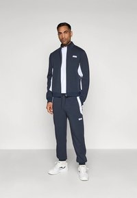 Granatowy dres wykonany z miękkiej tkaniny; rozpinana bluza z białymi panelami po bokach, w komplecie z dopasowanymi spodniami. Bluza zdobiona jest logo.