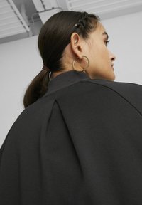 Schwarzer Sweatshirt mit hohem Kragen, mit einer Falten-Detail auf der Rückseite und gerippten Manschetten. Getragen mit einem großen Creolenohrring.