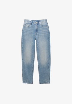Persoon die een grijze off-shoulder trui, lichtblauwe hoge taille jeans, zwarte sokken en bruine veterschoenen draagt, staand tegen een witte muur.