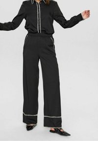 Chemise noire à manches longues avec bordure blanche contrastante le long du col et de la patte de boutonnage, associée à un pantalon noir à jambes larges avec coutures décoratives.