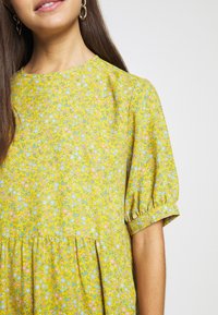 Keltainen kukallinen mekko, jossa on lyhyet puff-hihat, ja johon on kirjailtu vaaleanpunaisia ja sinisiä kukkakuvioita. Sujuva kangas, koottu vyötärödetail, rento malli.