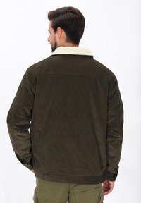 Jachetă din catifea în verde închis, cu un guler din material pufos de culoare crem. Are o cusătură pe spate și manșete cu nasturi. Suprafață texturată cu dungi verticale.