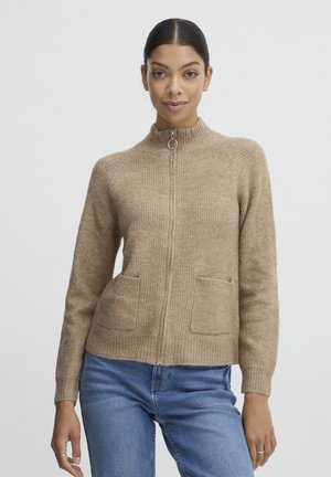 Cardigan lavorato a maglia beige con collo alto, chiusura a zip e due tasche frontali. Presenta una texture a costine e polsini aderenti.