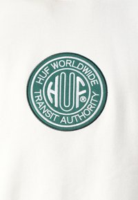 Tessuto bianco con una toppa rotonda ricamata, con testo verde e bianco che recita "HUF WORLDWIDE TRANSIT AUTHORITY" e un logo centrale.
