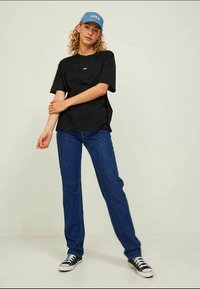 Tricou oversized negru, blugi albaștri și adidași negri cu accente albe. Modelul poartă o șapcă bleu deschis cu text. Design simplu, potrivire casual.