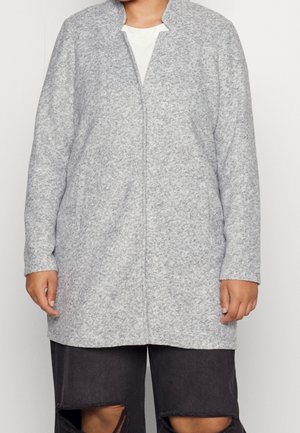 Person iført en lysegrå, tekstureret cardigan med lange ærmer over en hvid top, kombineret med sorte bukser med store knærevner.