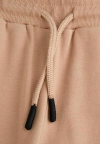 Beżowe spodnie dresowe z ryflowanym paskiem w talii, z plecionym sznurkiem do regulacji zakończonym czarnymi końcówkami. Gładka faktura, minimalizm w elementach designu.